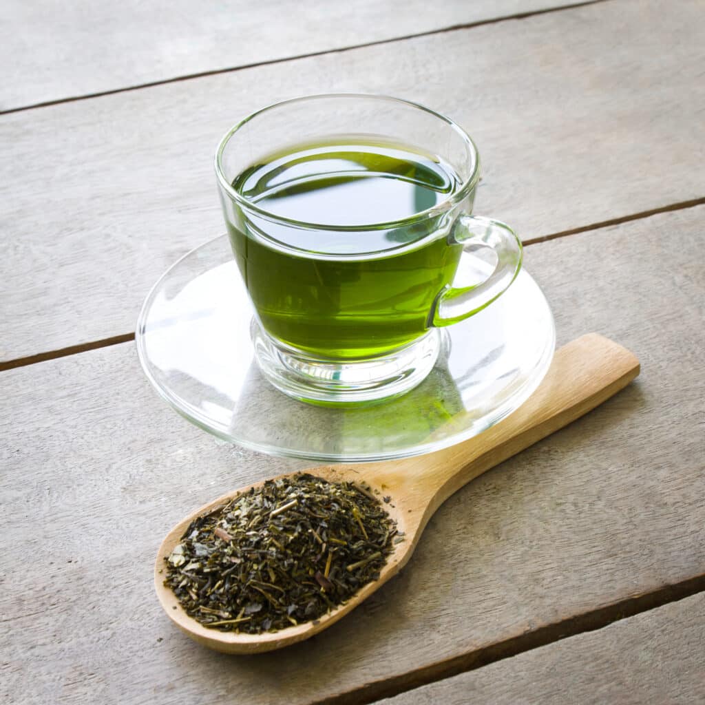 A cup of hot green tea and wooden spoon of dried green tea leaves on wooden base.